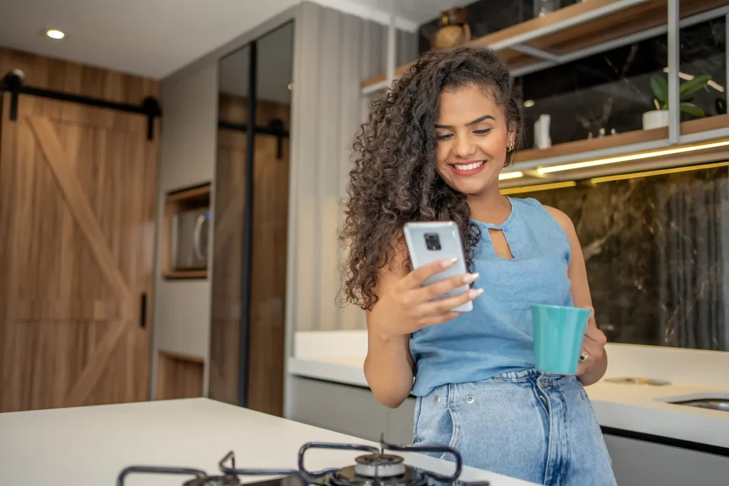 Mulher em uma cozinha com celular e caneca de café na mão